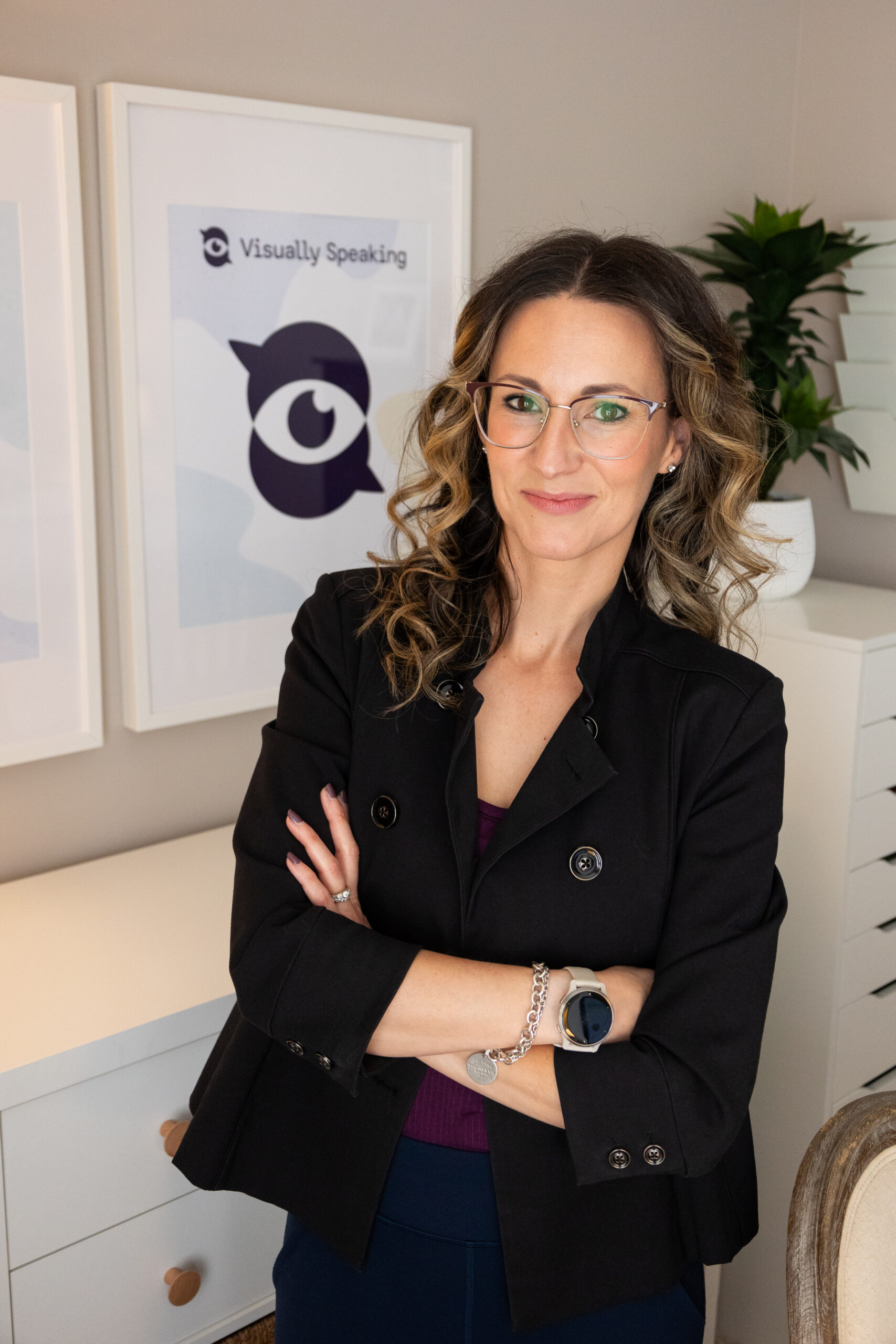 Professional portrait of Melissa Choquette standing with arms folded in a bright workspace, wearing glasses and a dark blazer, with a framed Visually Speaking logo displayed behind her.